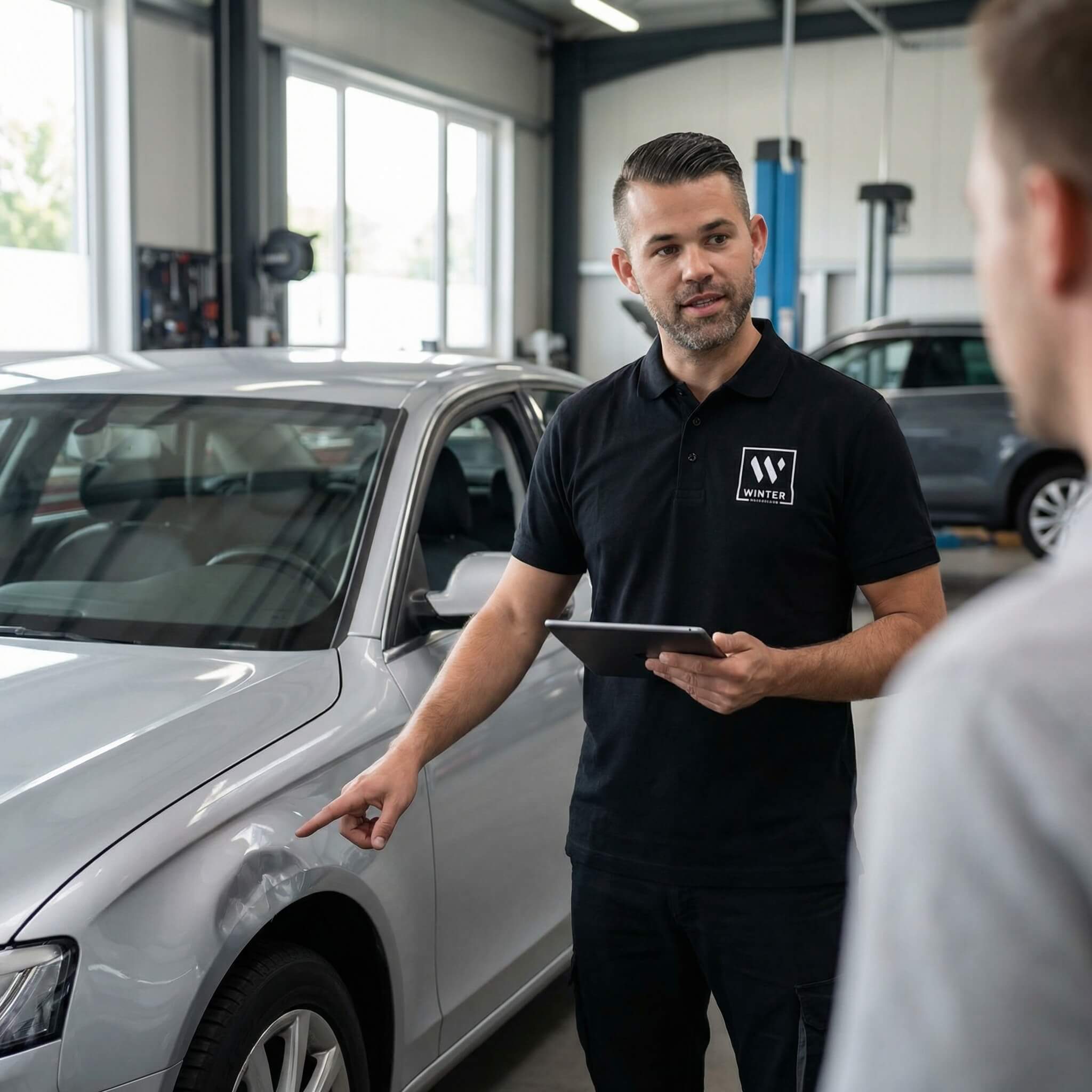 Ein Mann im schwarzen Poloshirt zeigt auf eine Delle am silbernen Auto und bespricht das Unfallgutachten mit einer weiteren Person