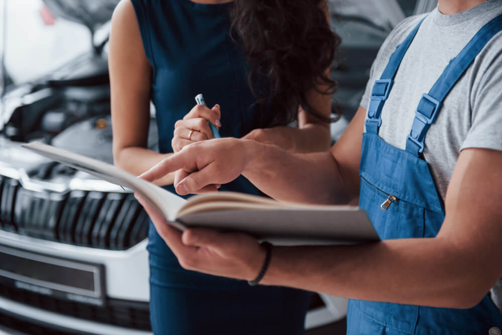 Ein Mechaniker in blauer Arbeitskleidung bespricht mit einer Frau vor einer geöffneten Motorhaube ein Dokument in der Autowerkstatt