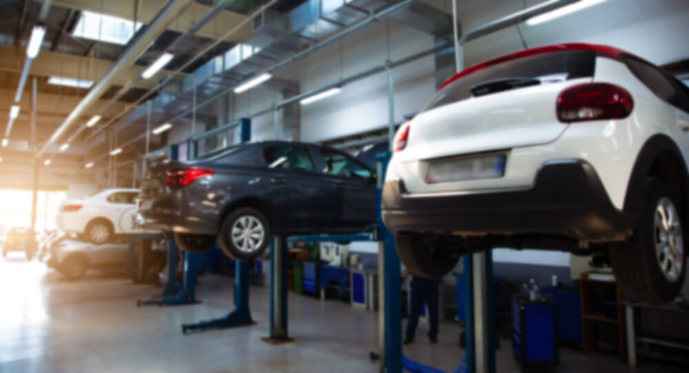 Mehrere unscharfe Autos stehen auf Hebebühnen in einer hell erleuchteten Autowerkstatt mit sichtbaren Arbeitsplätzen